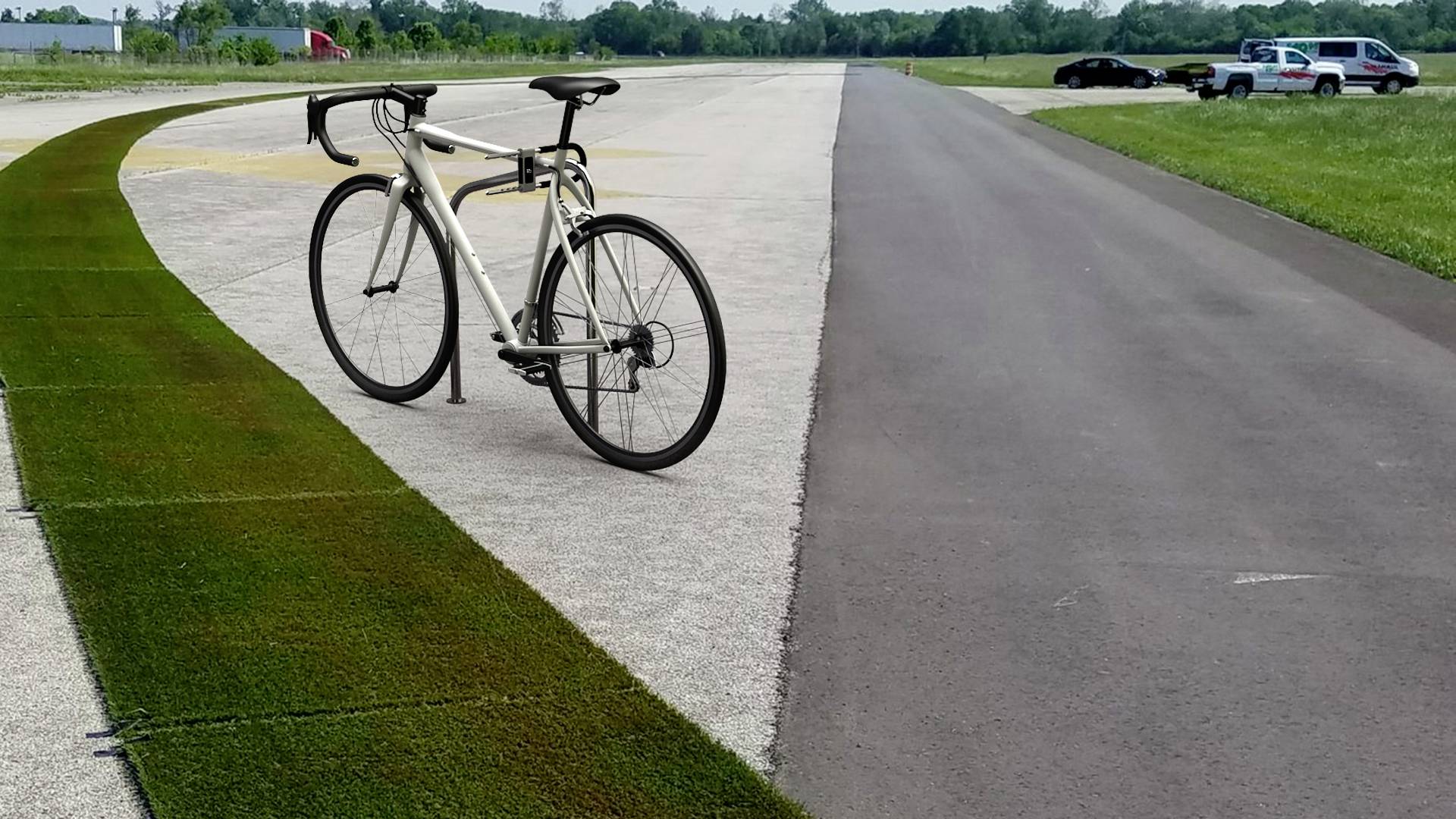 Silniční kolo stojí na široké dlážděné cestě vedle asfaltované silnice, obklopené poli a stromy v pozadí.