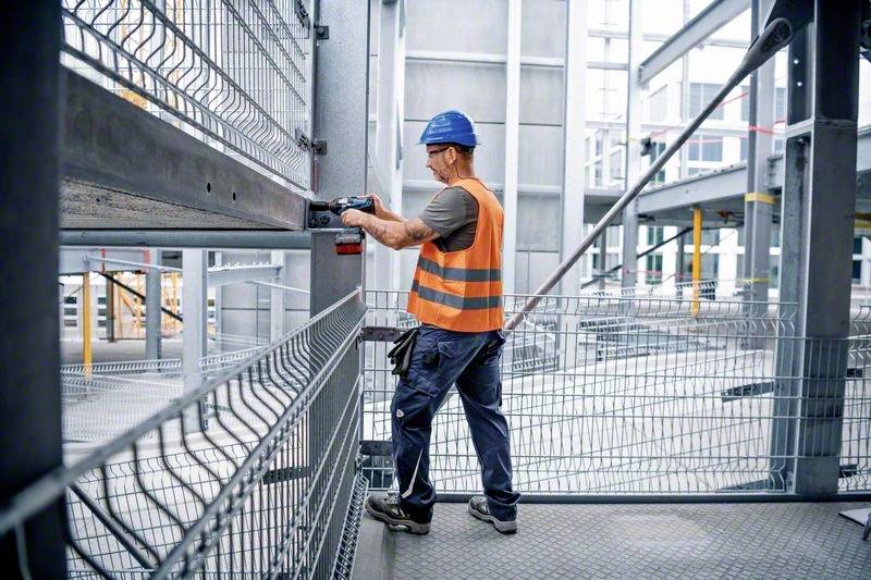 Dělník v oranžové vestě a modré přilbě upevňuje kovový nosník pomocí vrtačky na staveništi.