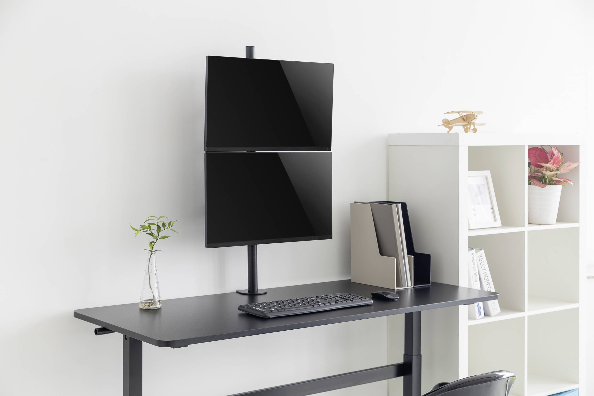 Elegantní výškově nastavitelný pracovní stůl se dvěma vertikálními monitory, klávesnicí, myší a malým květináčem vedle na světlém pozadí.
