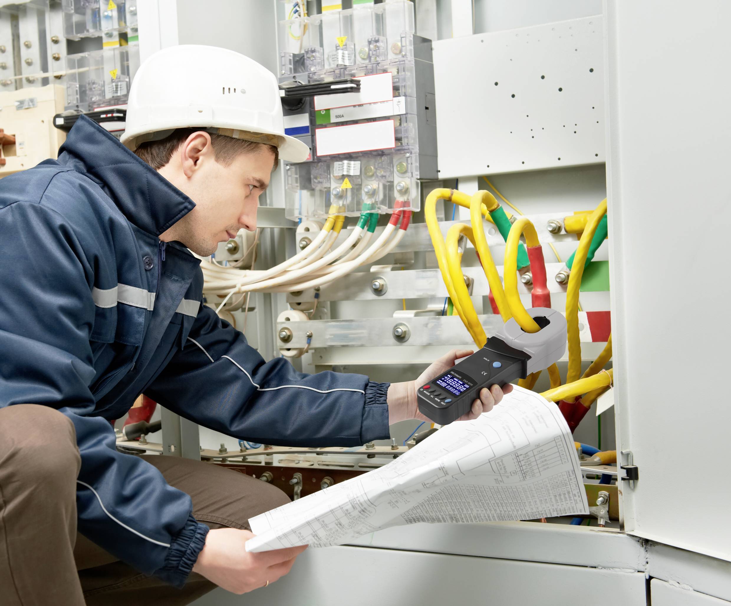Technik v ochranné přilbě kontroluje elektrické spoje v rozvaděči. Drží měřicí přístroj a stavební plány.