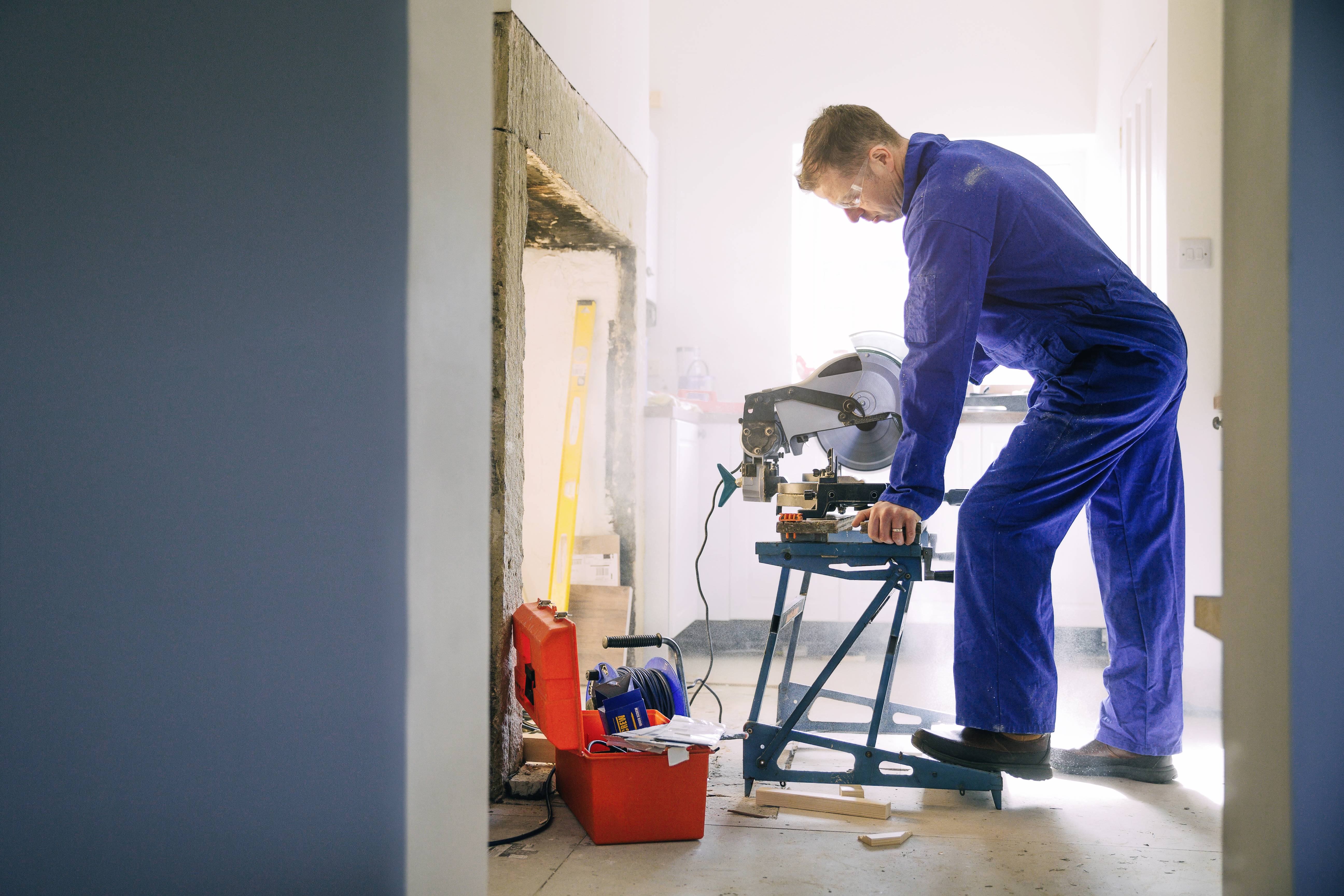 Muž v modrém kombinézonu soustředěně pracuje s pilou v světlé místnosti. Na podlaze leží bedna s nářadím a materiály.
