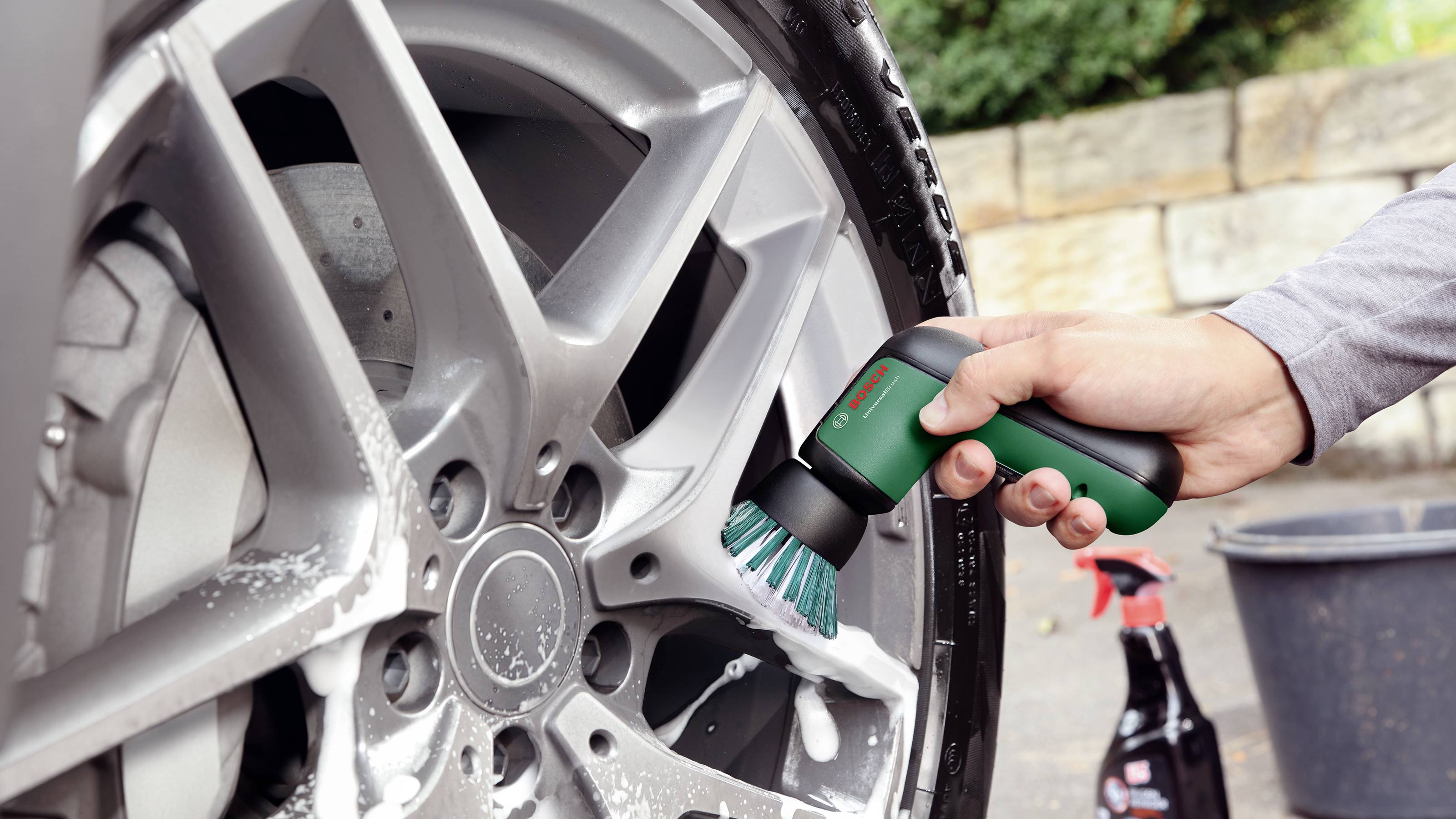 Nástroj čistí pěnou pokrytý ráfek automobilu. V pozadí stojí rozprašovač na zemi.