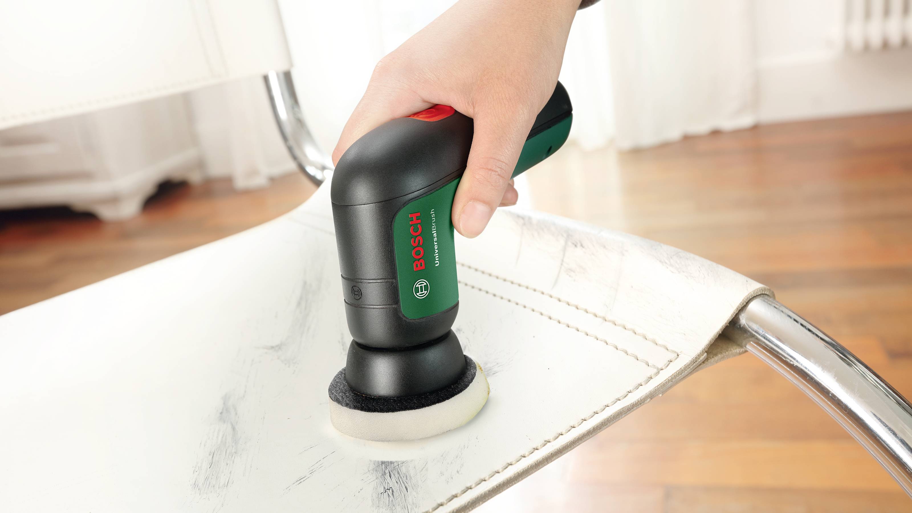 Jedna ruka drží elektrickou brusku a opracovává sedací plochu bílé židle na dřevěné podlaze.