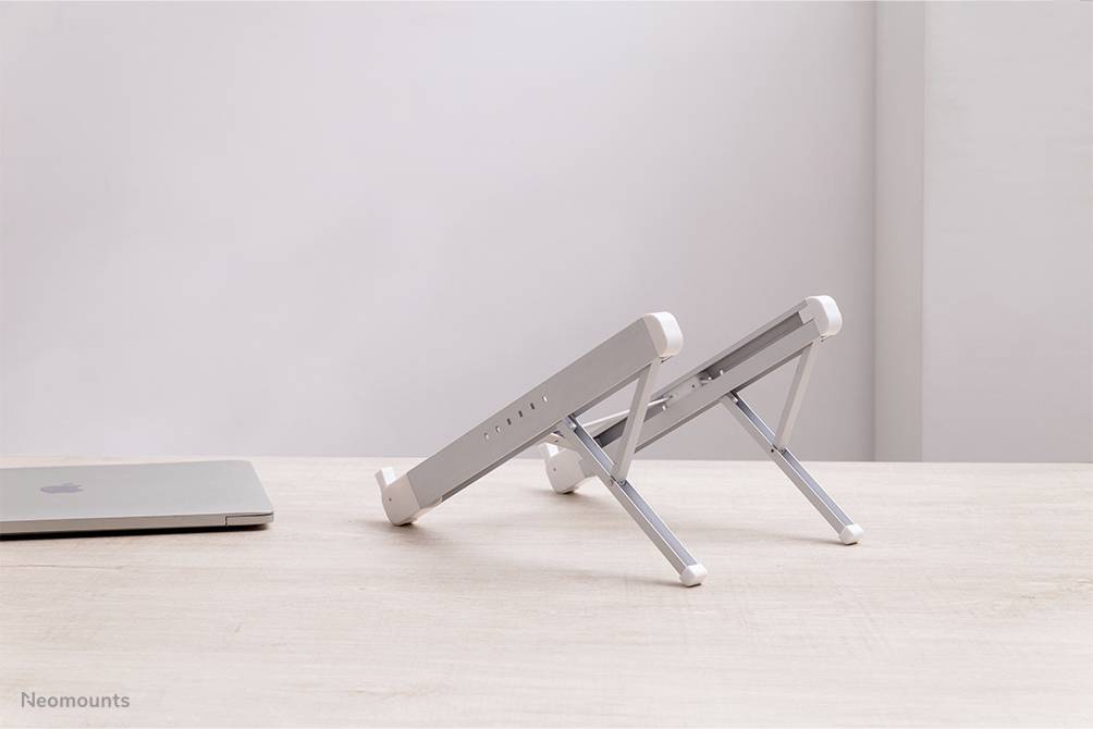 Stříbrný stojan na notebook na dřevěném stole vedle zavřeného laptopu. Pozadí je jednoduché a minimalistické.