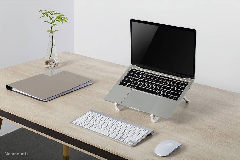 Laptop na stole stojí na stojanu, vedle něj klávesnice a myš. Vpravo sklenice s rostlinou, vlevo uzavřený notes.