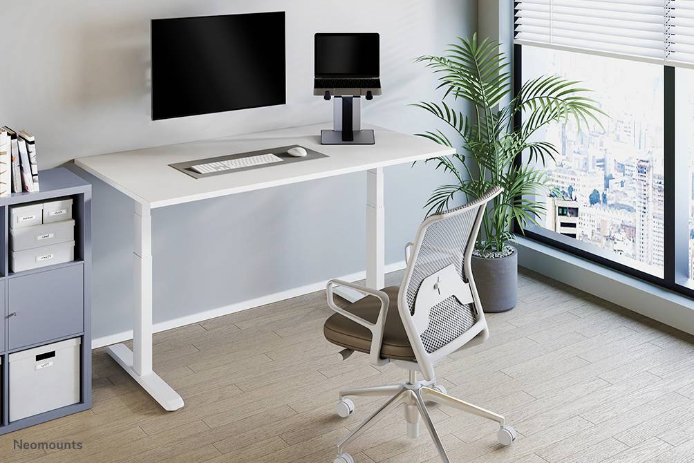 Výškově nastavitelný psací stůl s počítačem a ergonomickou židlí, vedle okna s rostlinami a výhledem na město.