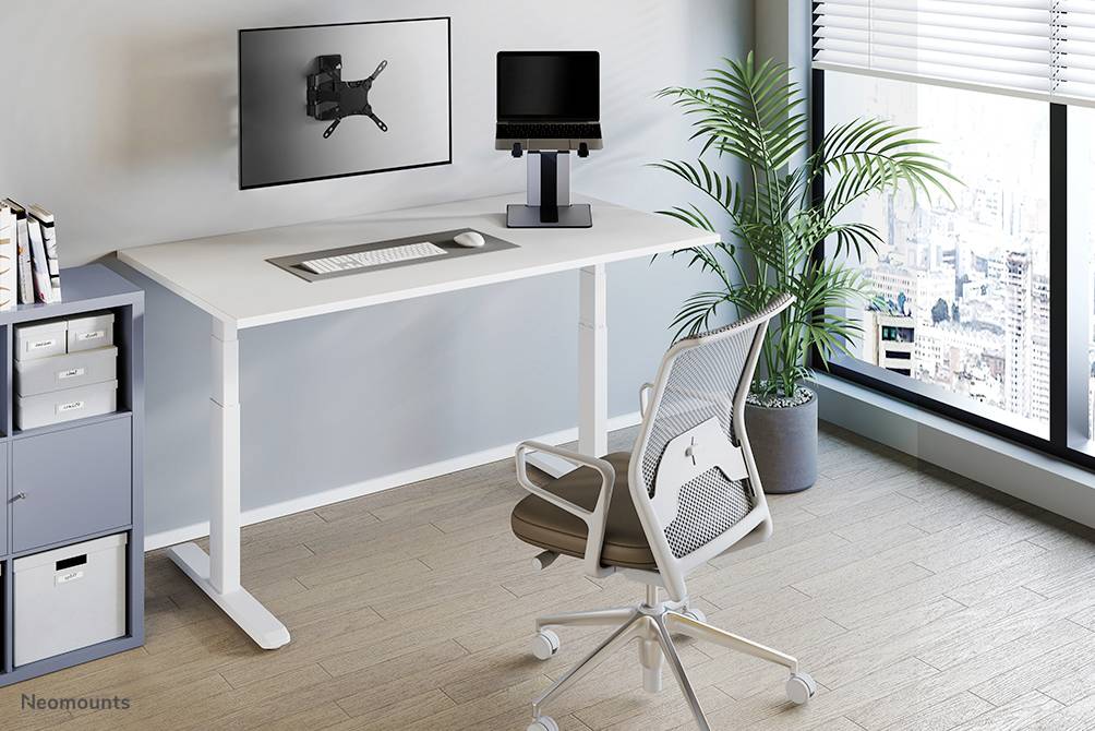 Výškově nastavitelný psací stůl s monitorem na držáku, klávesnicí, stojanem na laptop na stole, moderní kancelářské prostředí s oknem.