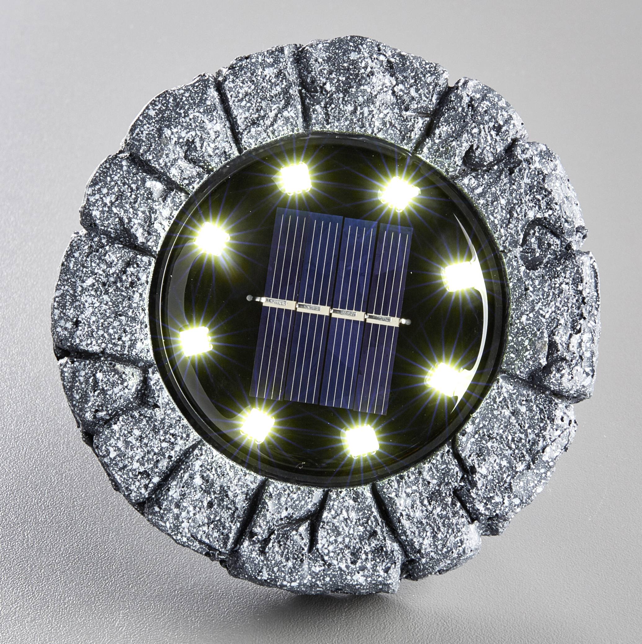 Kulatý, kamenu podobný solární reflektor s osmi svítícími LED diodami kolem centrálního solárního panelu na šedém povrchu.