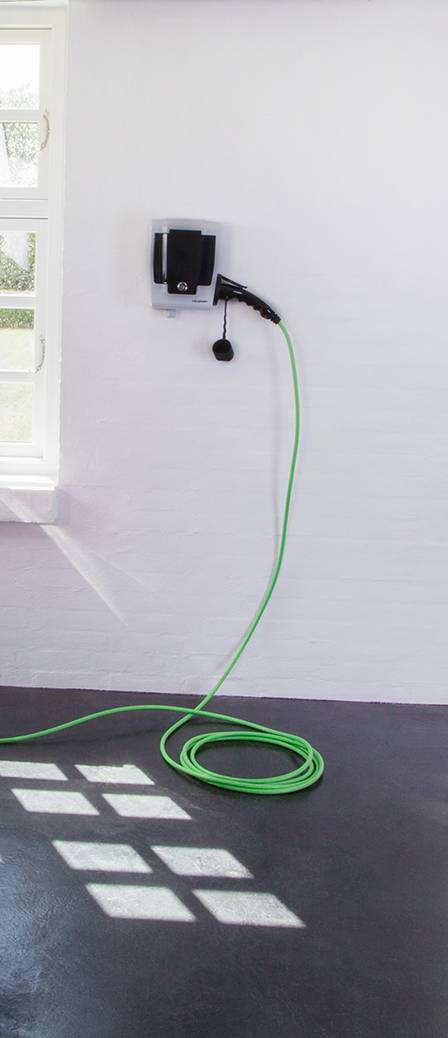 Nabíjecí kabel pro elektromobily se zeleným kabelem visí na bílé stěně, pod ním je okno, které vrhá světlo na podlahu.