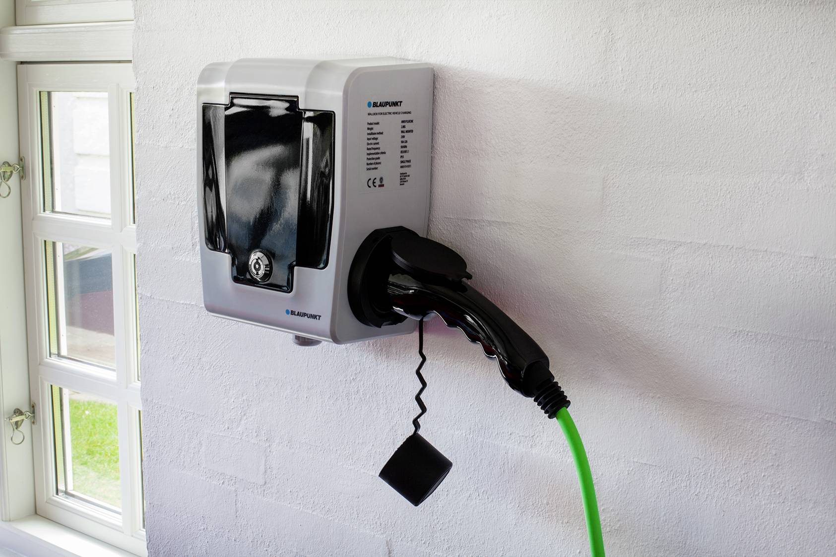 Nabíjecí stanice pro elektromobily s připojeným nabíjecím kabelem u bílé stěny. Vpravo v obraze je okno.