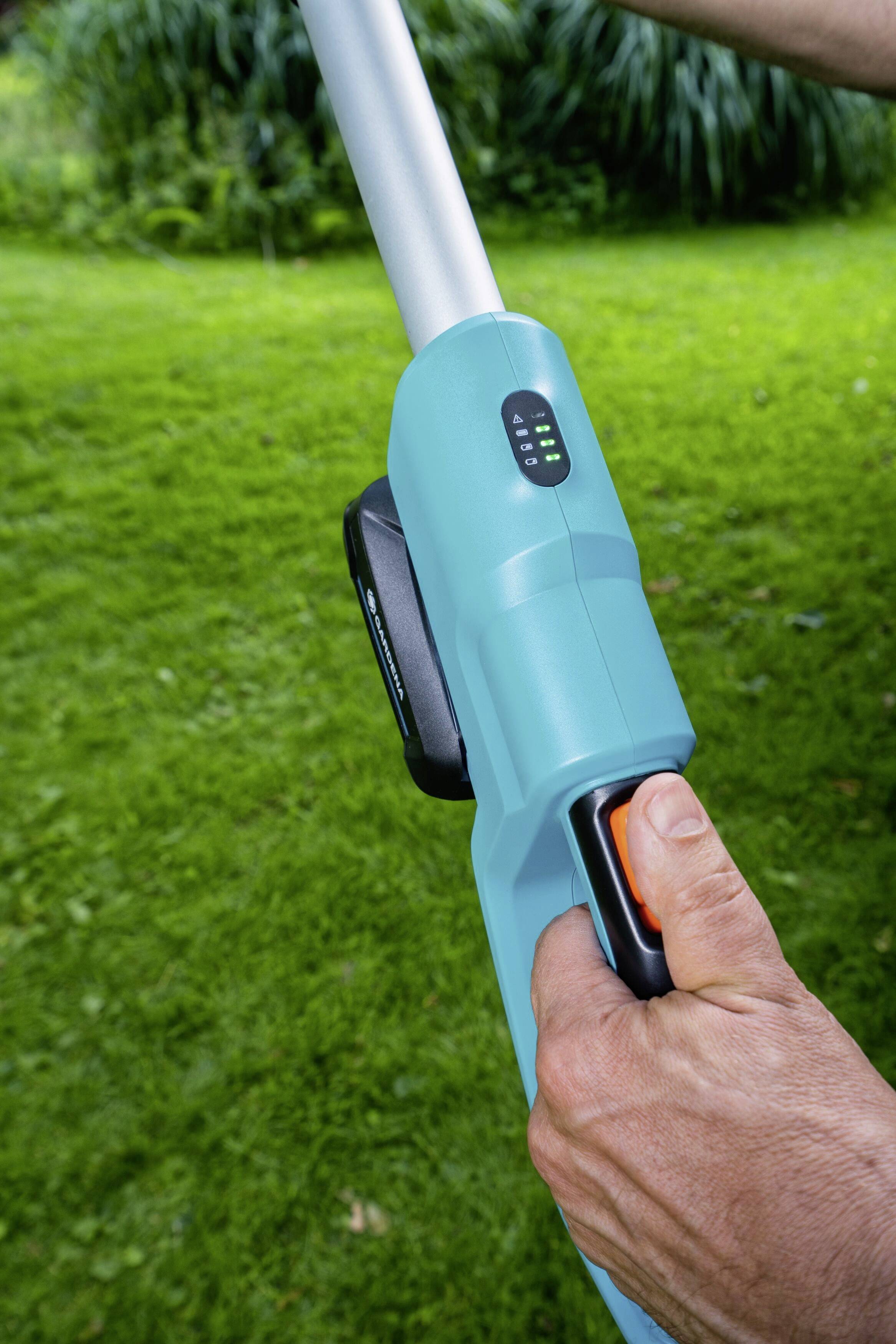 Osoba drží modrý elektrický rukojeti zahradního nástroje s černým spínačem na zeleném trávníkovém pozadí.