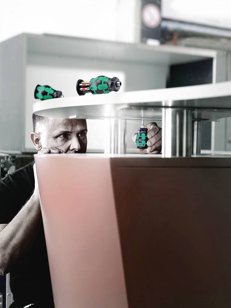 Muž pečlivě montuje kovovou součástku v dílně s černo-zelenými šroubováky. Soustředí se na preciznost.