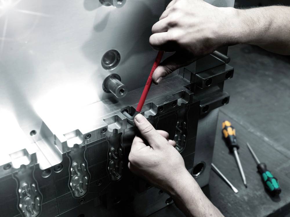 Někdo seřizuje kovový stroj červeným šroubovákem. V pozadí leží další dva šroubováky.