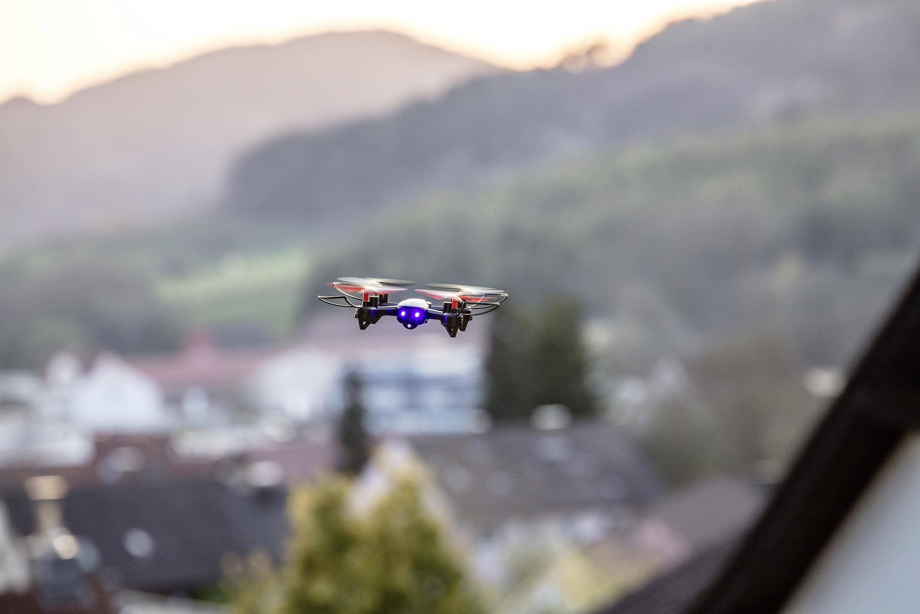 Dron se vznáší nad městskou krajinou při západu slunce. V pozadí jsou rozmazané kopce a domy.