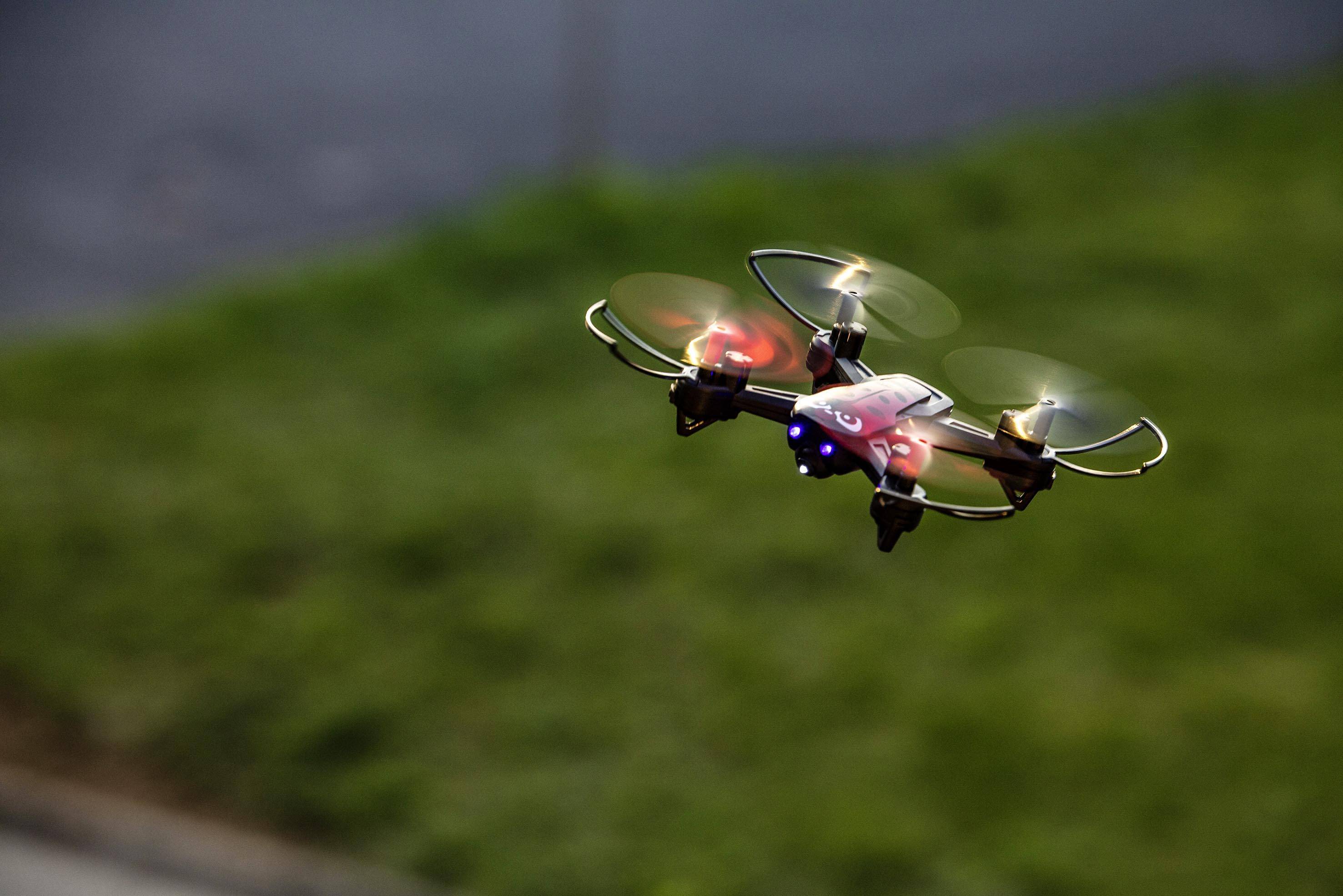 Dron se zářícími vrtulemi letí v nízké výšce nad rozostřenou zelenou loukou.