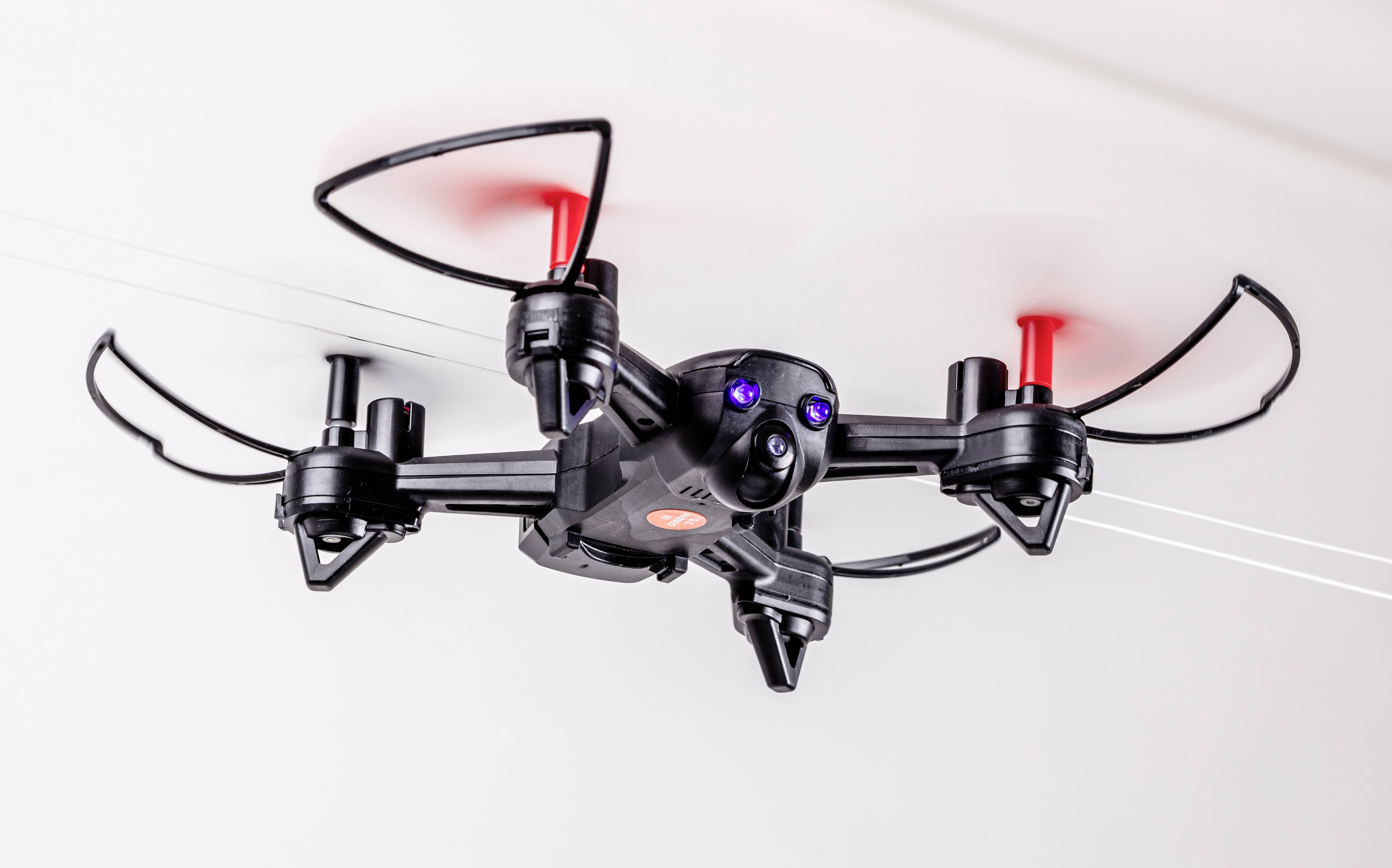 Černý dron s červenými vrtulemi letí vnitřním prostorem. Dron je vybaven kamerou a nachází se v pohybu.