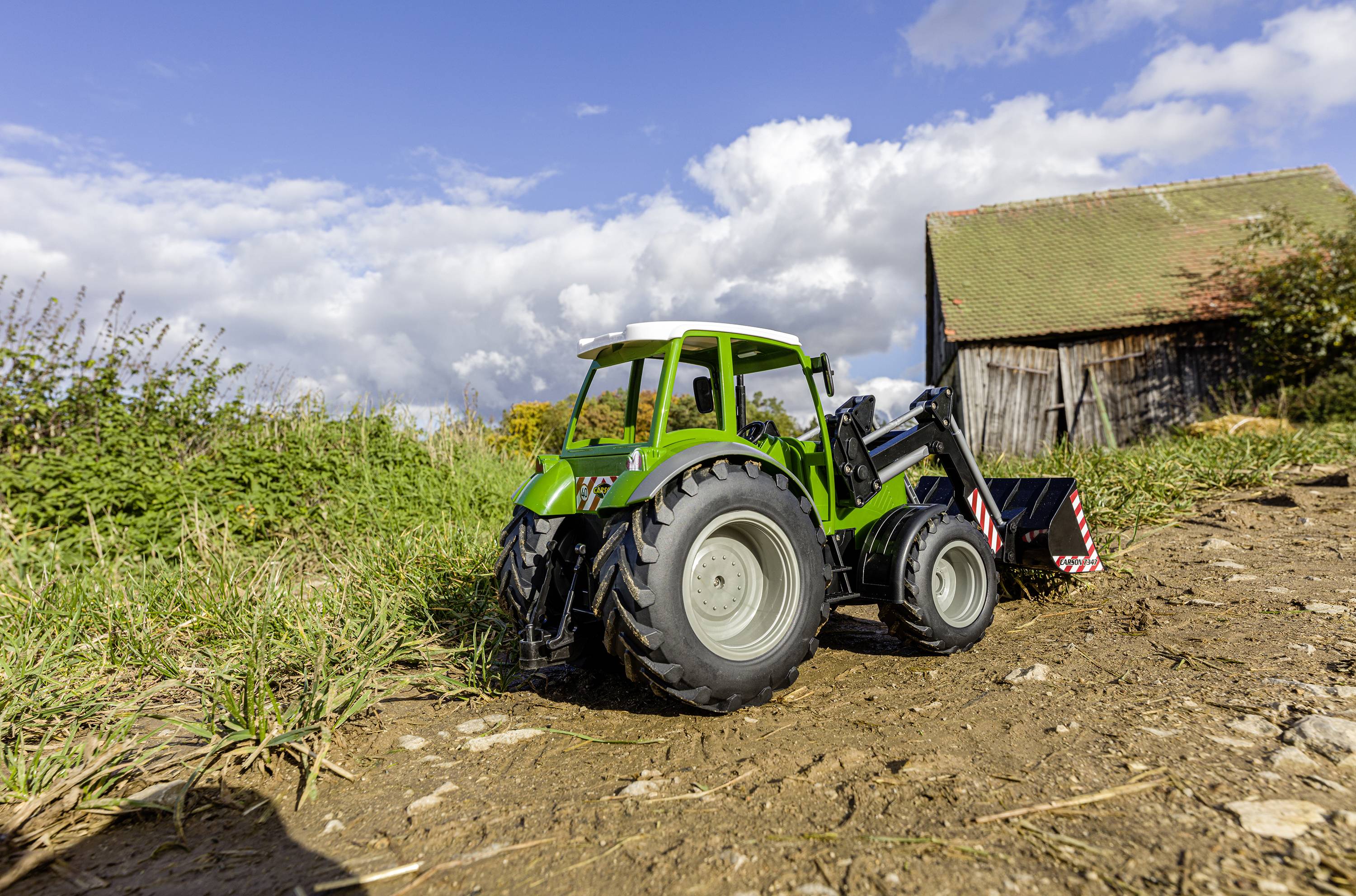 Zelený hračkářský traktor na polní cestě před kulisou připomínající stodolu pod modrou oblohou s mraky.