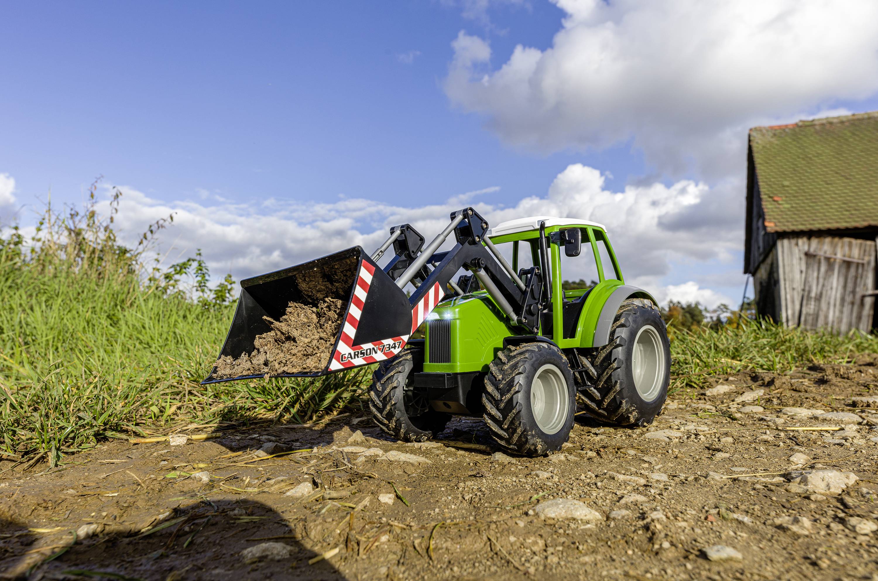 Zelený hračkářský traktor s lopatou přepravuje hlínu po polní cestě. V pozadí je vidět stodolu.