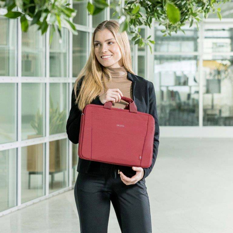Žena stojí v moderní kanceláři s červeným obalem na notebook v pravé ruce, obklopená rostlinami a skleněnými stěnami.