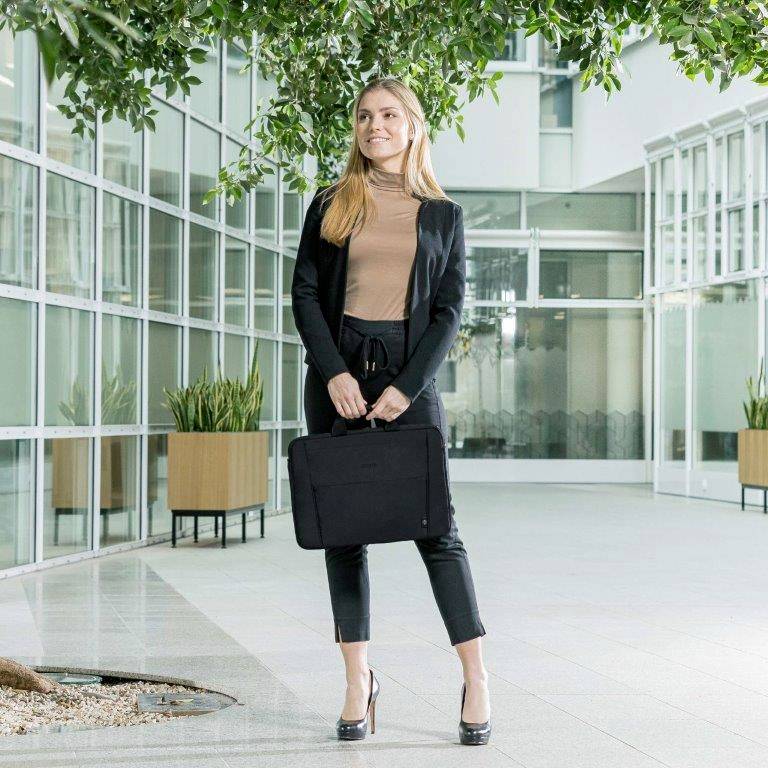 Žena stojí v moderní chodbě budovy s rostlinami, drží černou aktovku a usmívá se.