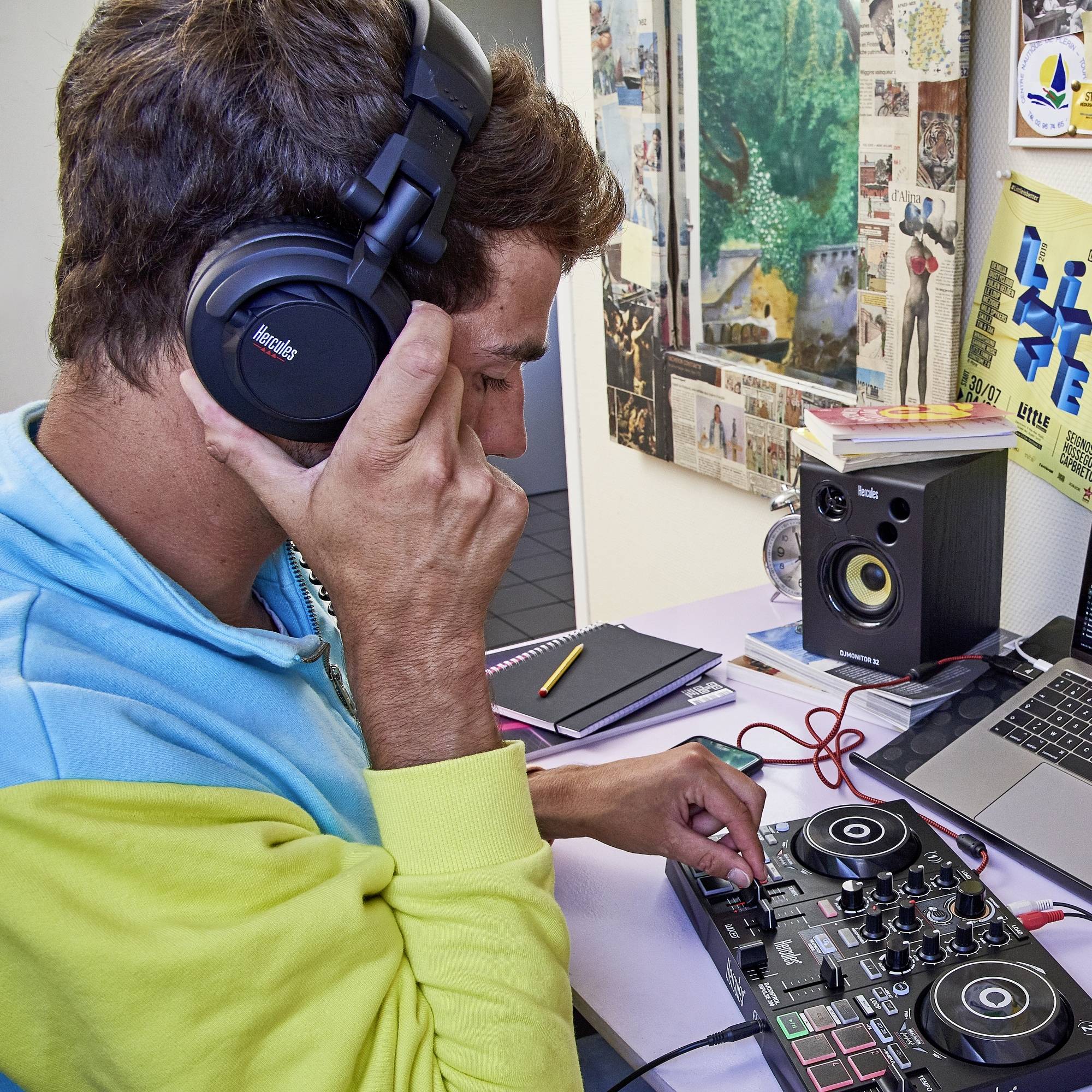 Osoba má sluchátka a ovládá DJ kontroler v místnosti s laptopem a reproduktory, obklopena plakáty a poznámkami.