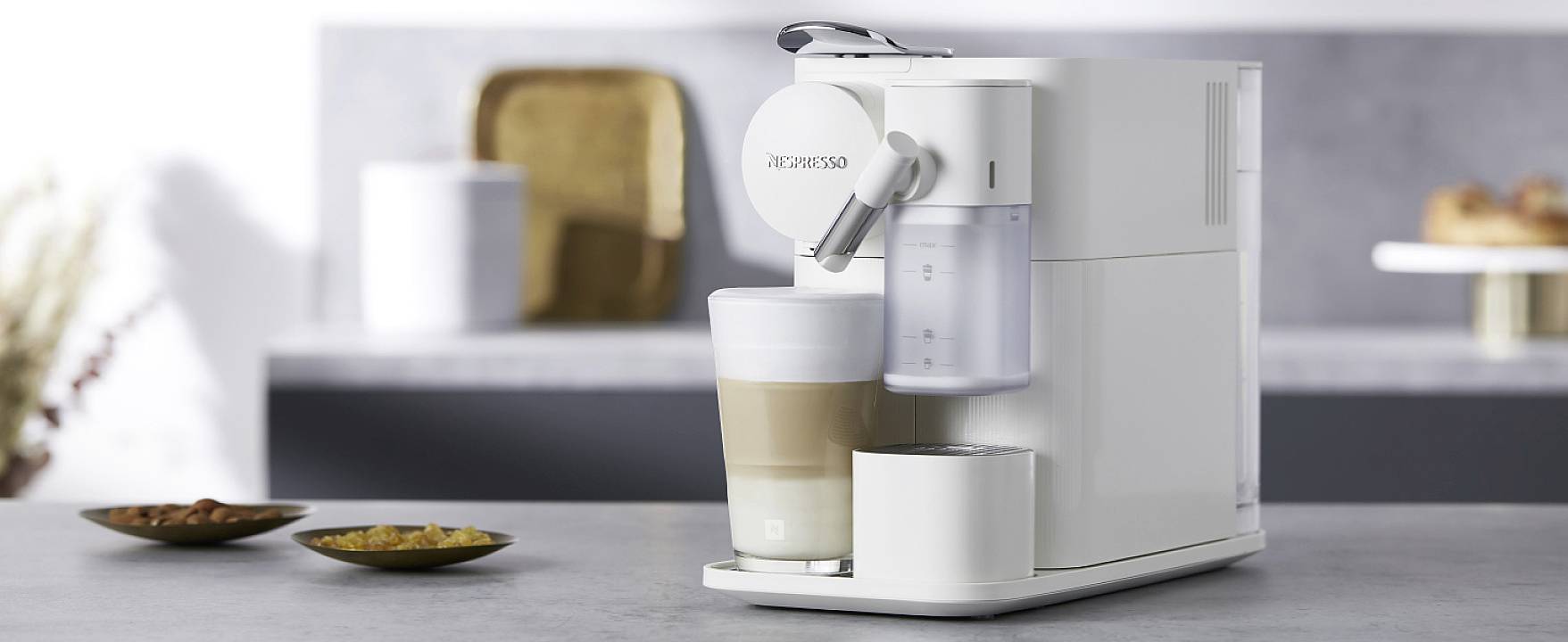 Bílý kávovar na šedé pracovní desce připravuje cappuccino. V pozadí rozmazané kuchyňské prvky a talíř.