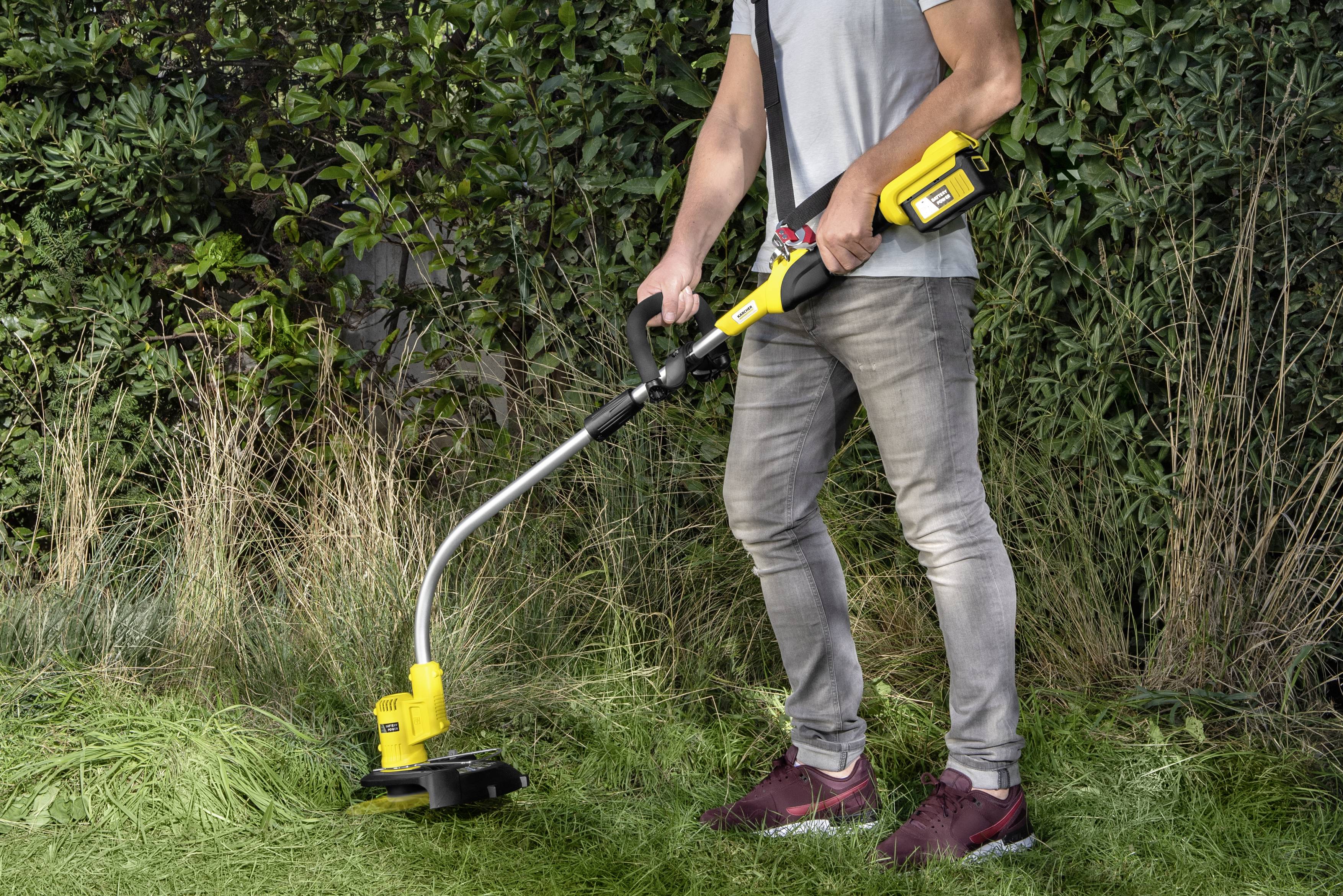 Osoba seká travní porost žlutým křovinořezem podél okraje zahrady. V pozadí jsou vidět keře.