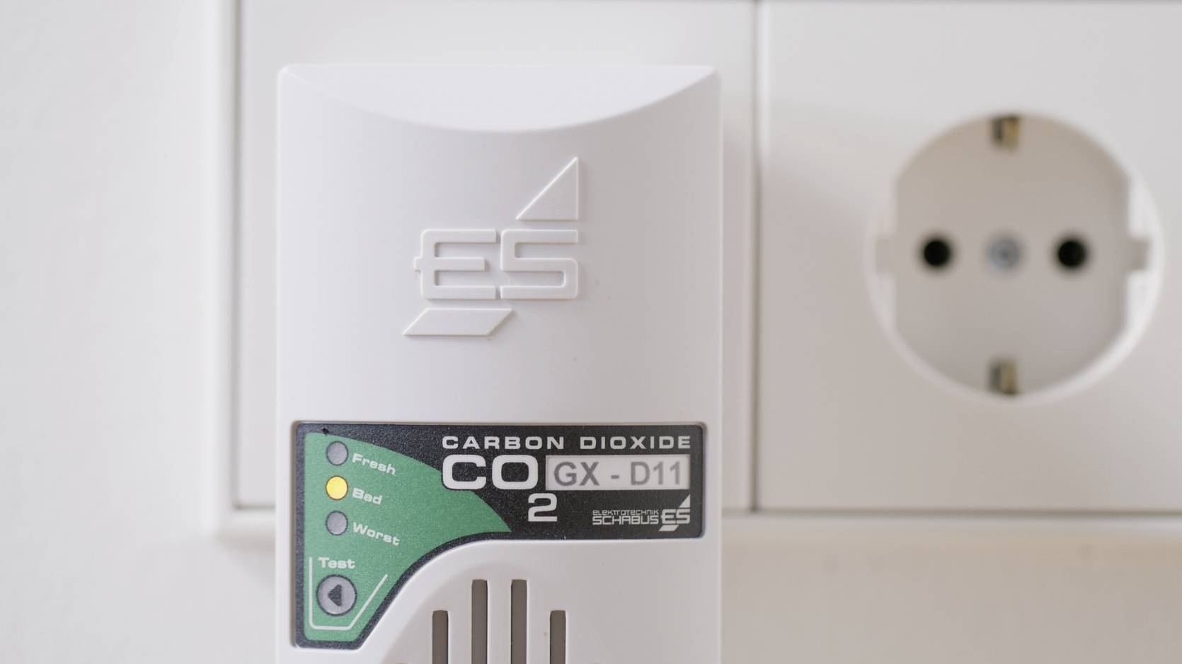 CO2 měřicí přístroj GS-D11 připojený do zásuvky. Na přední straně jsou indikátory stavu 'Čerstvý', 'Ideální', 'Varující' a 'Test'.