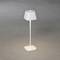 Bílá, minimalistická stolní lampa s čtvercovou základnou, stojící na šedé podlaze, svítící směrem nahoru.
