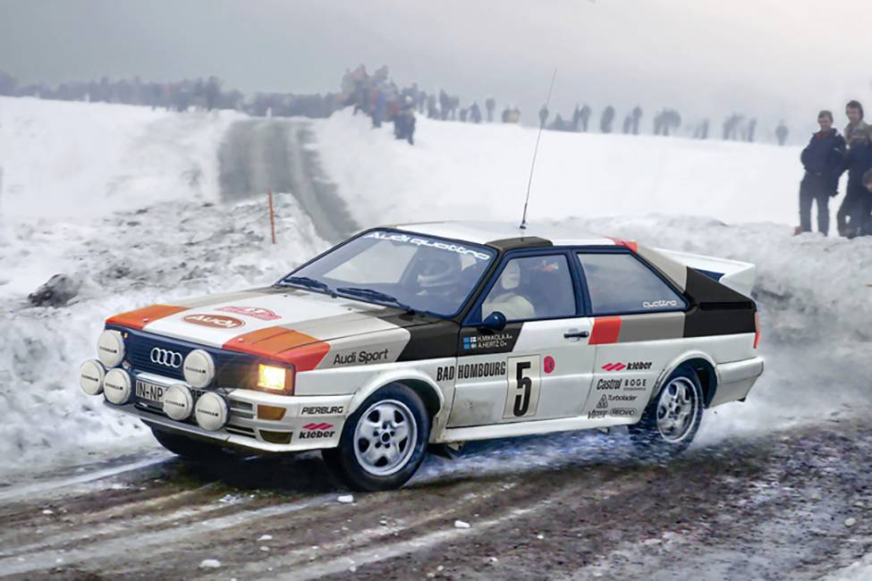 Rally auto jede rychle po zasněžené trati. Diváci stojí podél silnice. Auto má startovní číslo 5.
