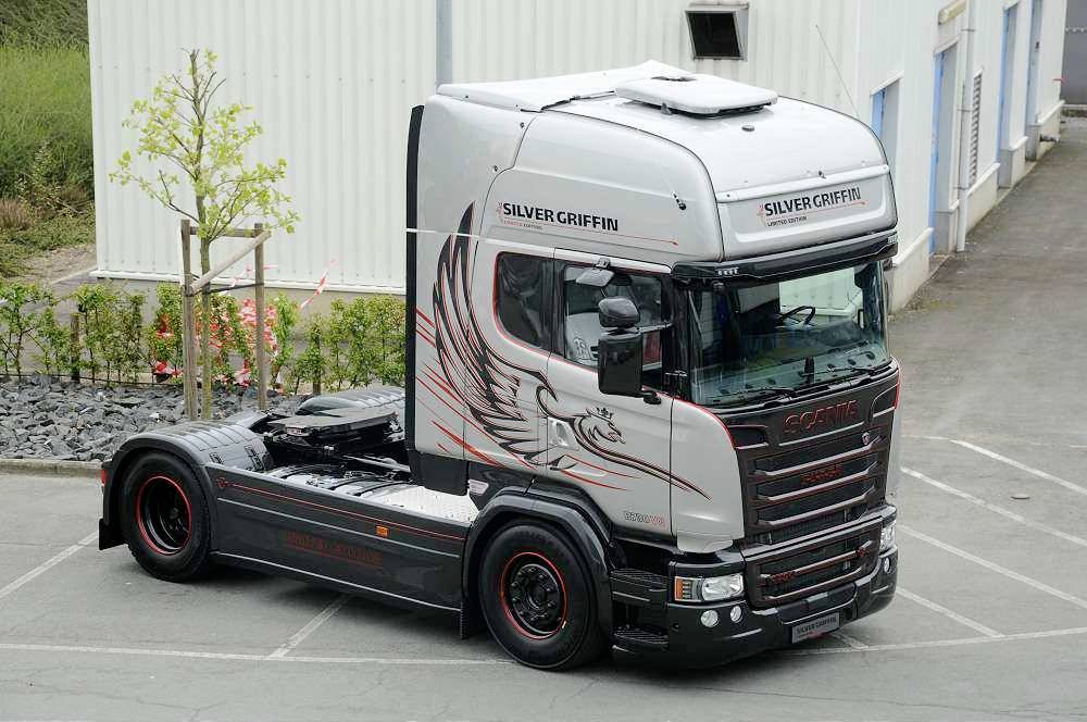 Stříbrný nákladní vůz značky Scania 'Silver Griffin' s orlí grafikou stojí na dvoře mezi budovami.