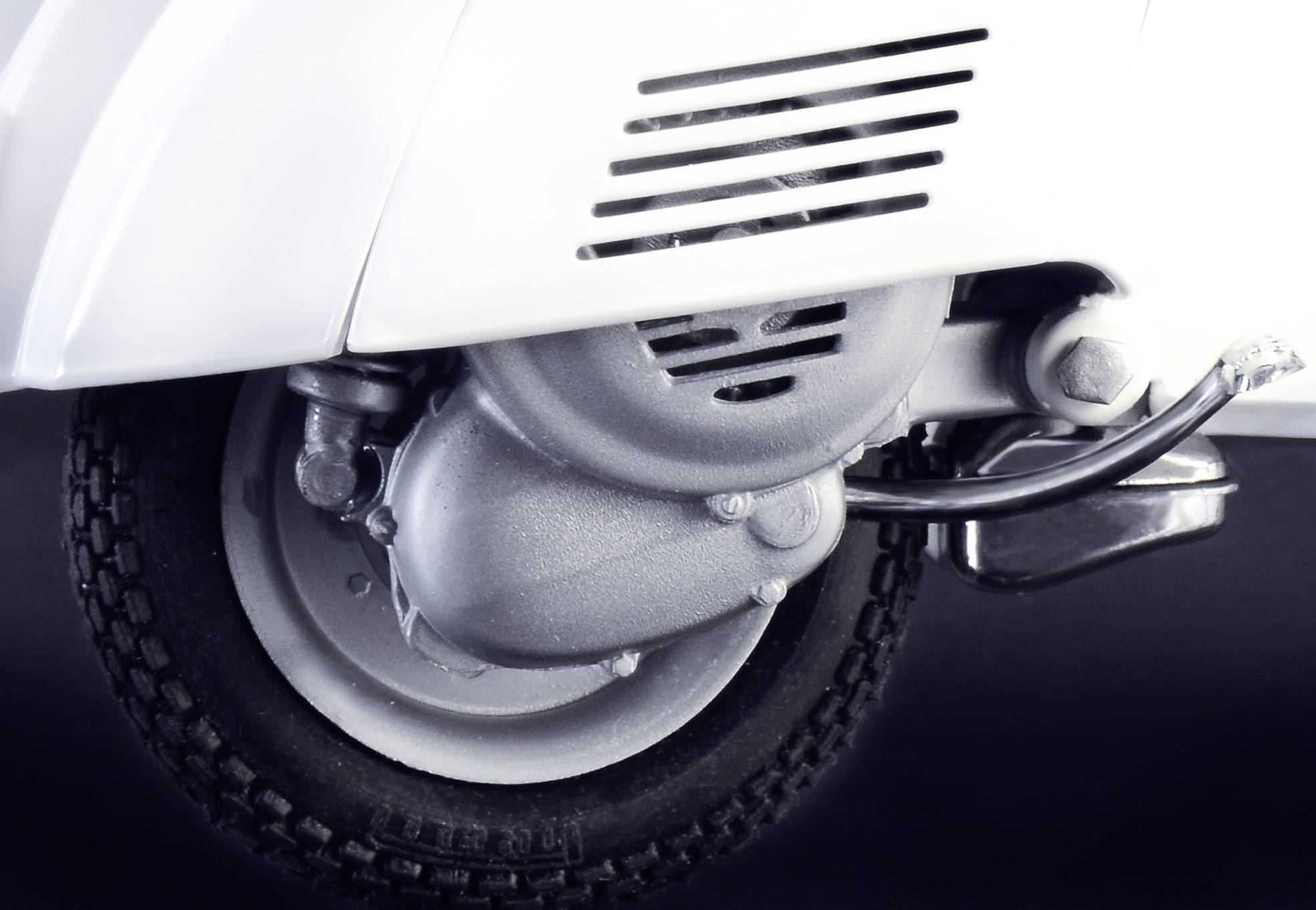 Detailní záběr bílého motorového skútru s viditelným motorem a větracími otvory na tmavém pozadí.