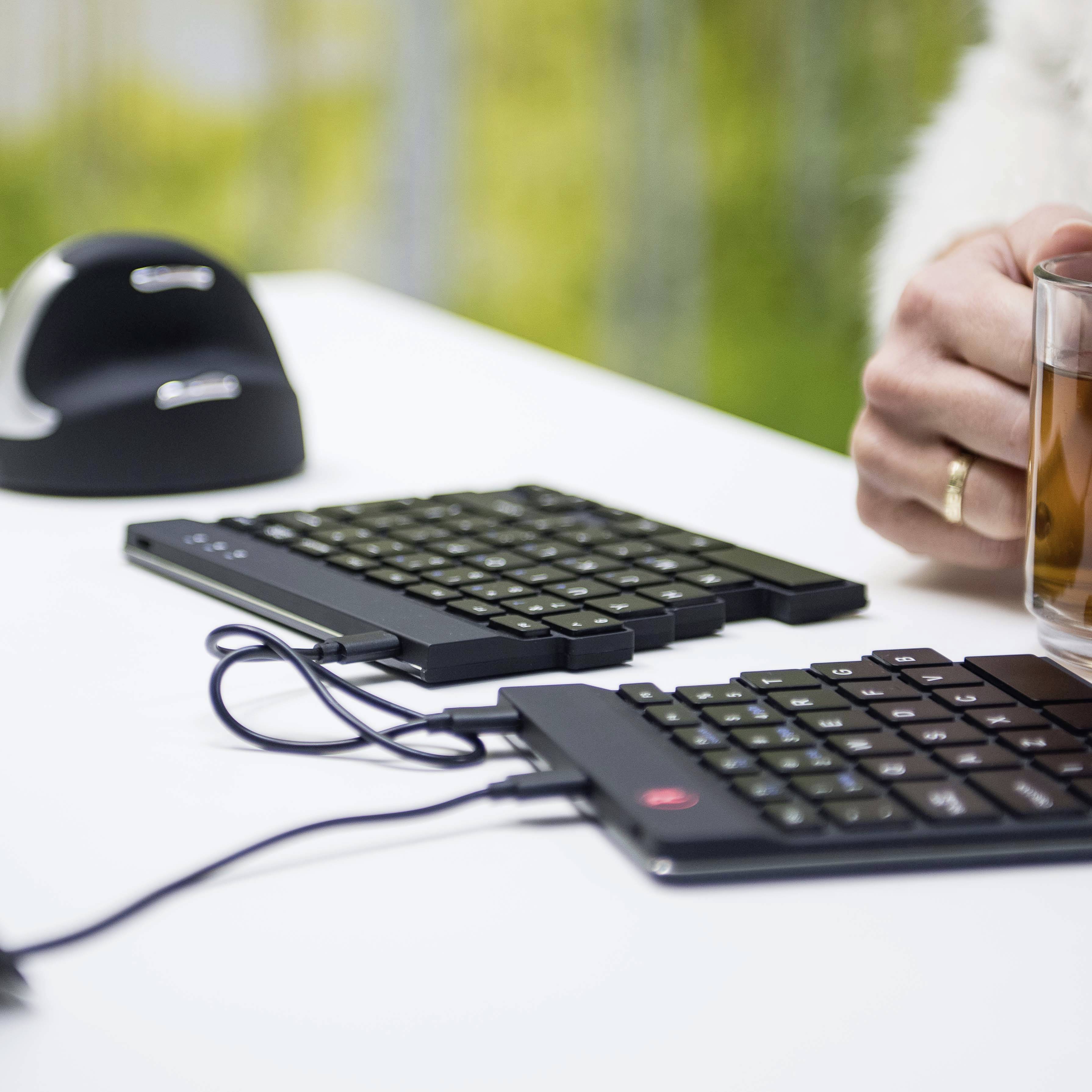 Jedna ruka drží sklenici čaje vedle ergonomické klávesnice na psacím stole. V pozadí rozostřená zelená vegetace.