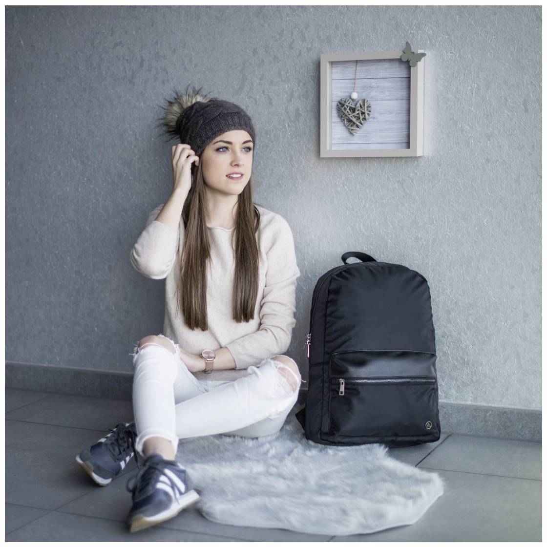 Žena sedí na podlaze v pohodlném oblečení, má na hlavě čepici, vedle ní leží černý batoh. Na stěně visí dekorace ve tvaru srdce.