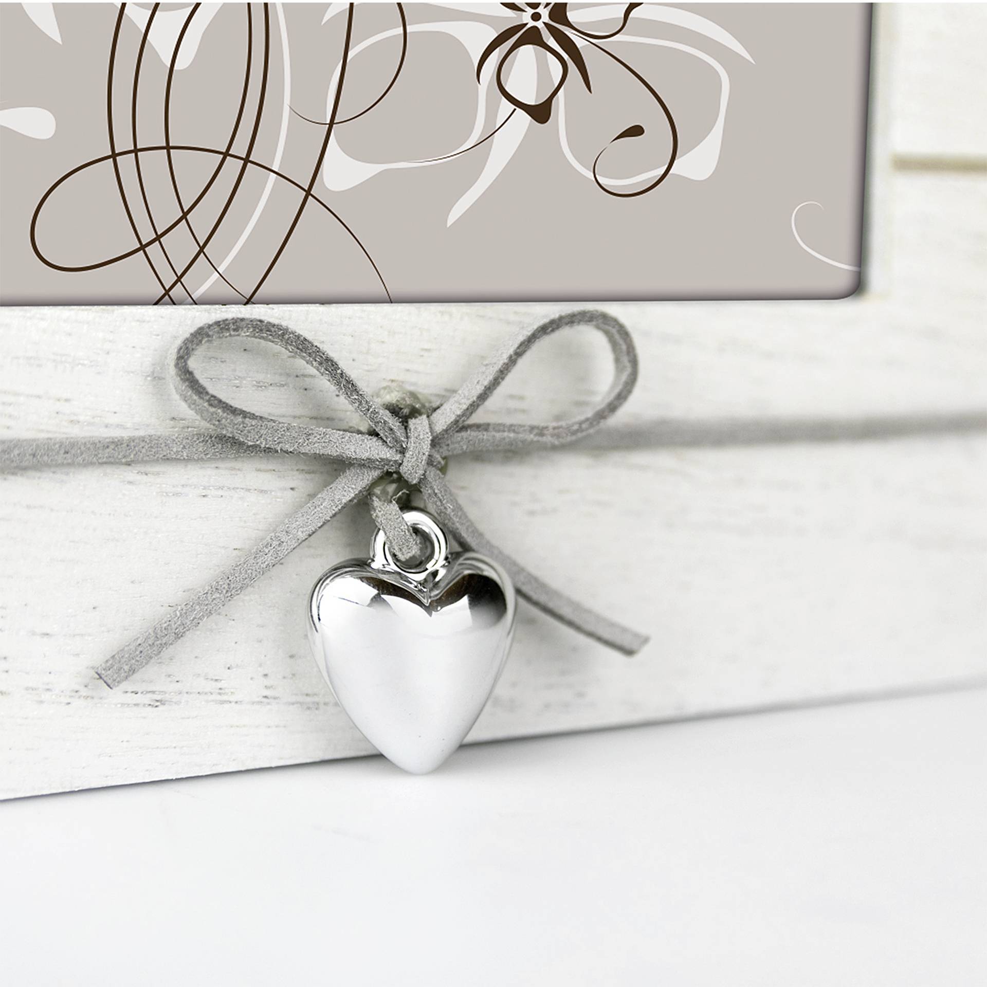 Stříbrný přívěsek ve tvaru srdce visí na šedé stužce před pozadím s abstraktními bílými květinovými vzory na béžovém podkladu.
