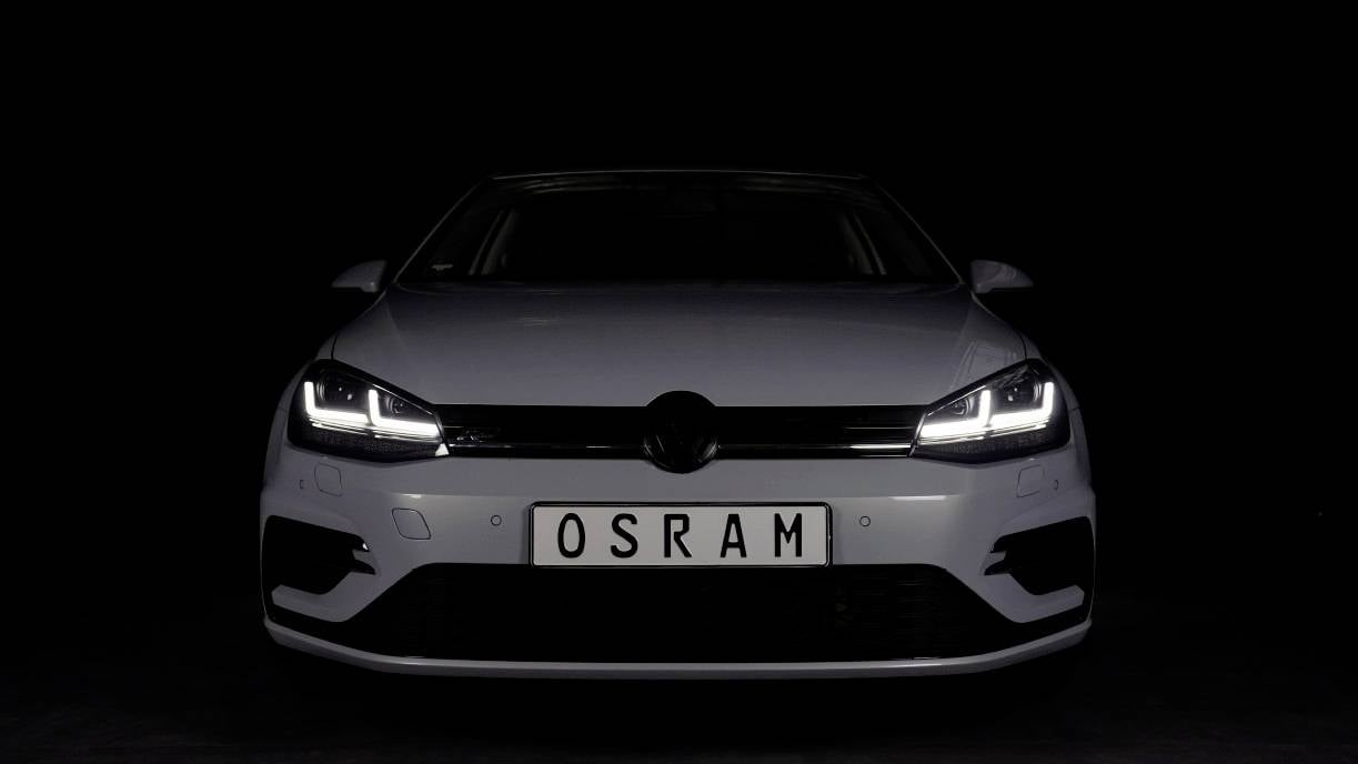 Bílé auto se zapnutým LED denním světlem, pohled zepředu na černém pozadí. Na poznávací značce je napsáno 'OSRAM'.