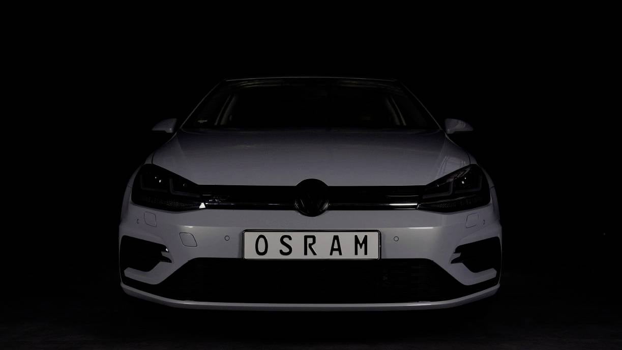 Bílé auto v tmavém prostoru s registrační značkou 'OSRAM', viditelné čelně. Důraz je kladen na osvětlení vozidla.