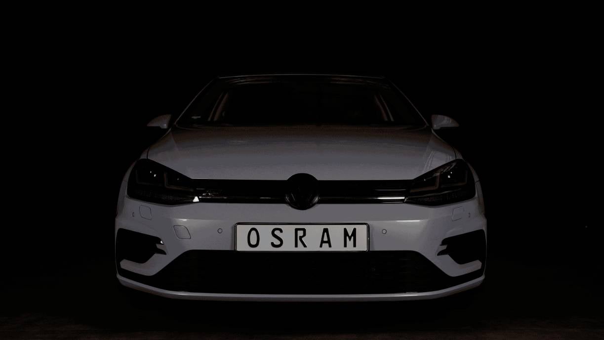 Auto se zapnutým světlometem v tmavém prostoru, s registrační značkou 'OSRAM', jehož světlomet výrazně vyčnívá na černém pozadí.