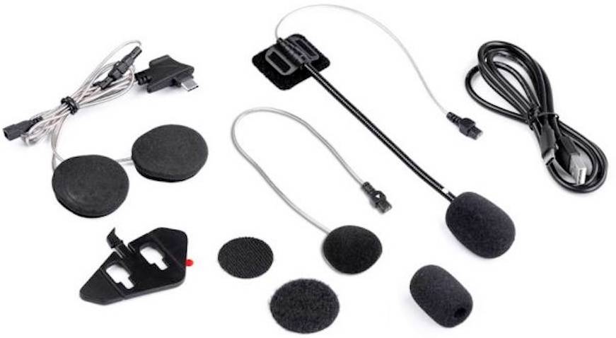 Sada černé motocyklové helmy a příslušenství pro headset, včetně mikrofonů, reproduktorů a připojovacích kabelů, umístěná na bílém pozadí.