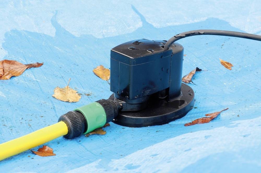 Černá vodní pumpa na modré plachtě, propojená žlutou hadicí. Několik podzimních listů je rozmístěno kolem pumpy.