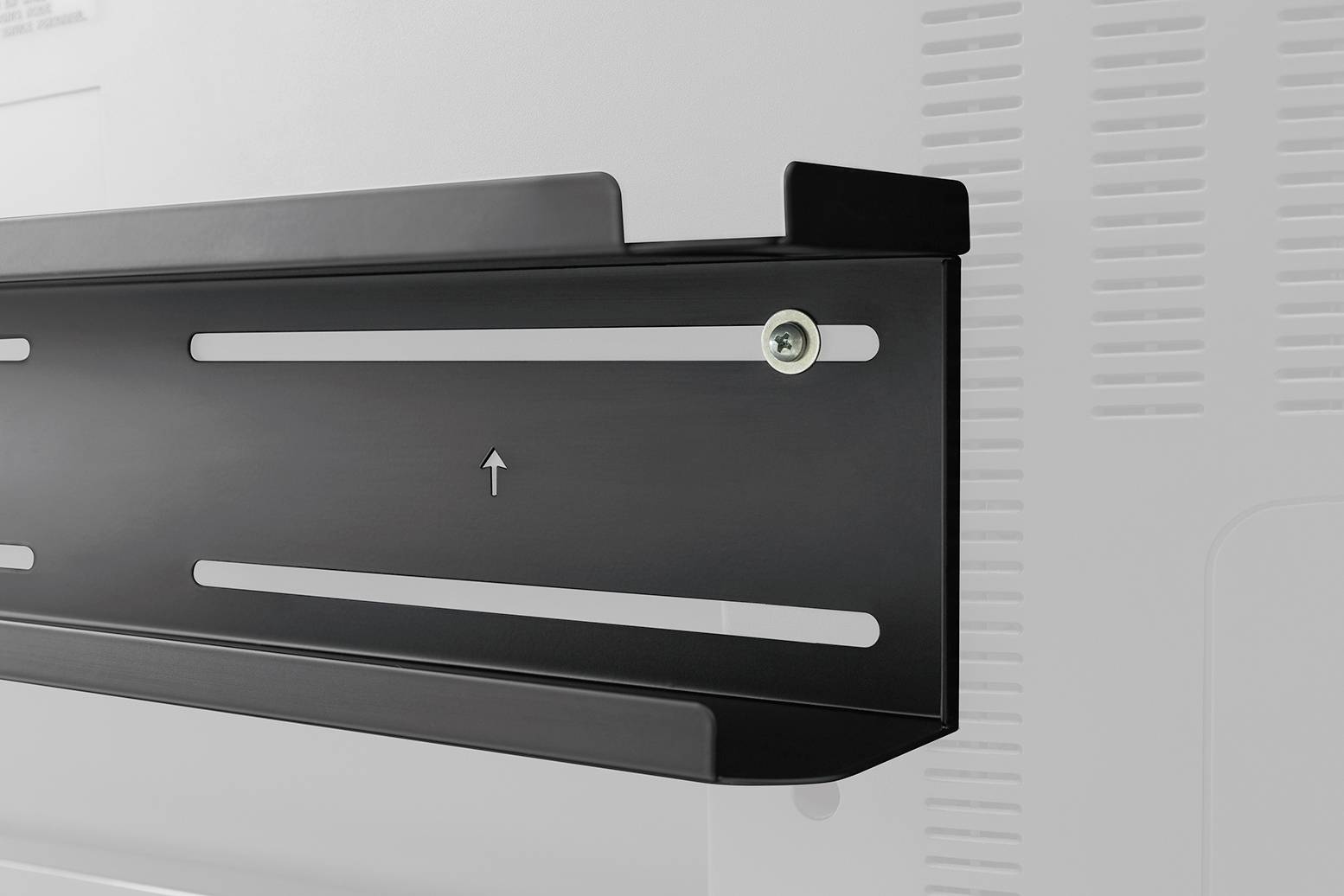 Detailní záběr černého nástěnného držáku se šroubem připevněného na bílé stěně; šipka směřuje nahoru, pravděpodobně pro montáž televize.