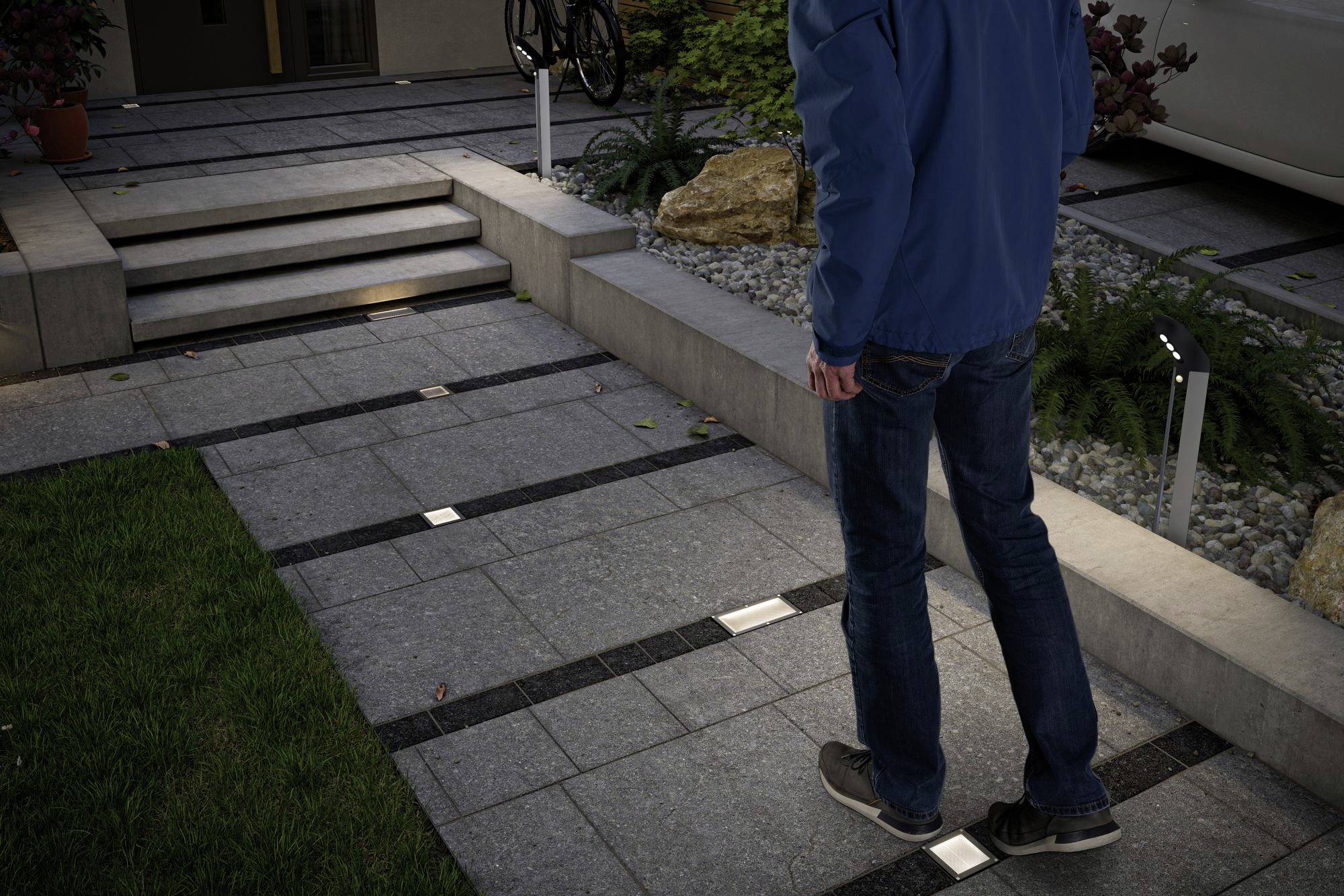 Osoba kráčí osvětlenou zahradní cestou s moderní dlažbou a schodištěm za soumraku.