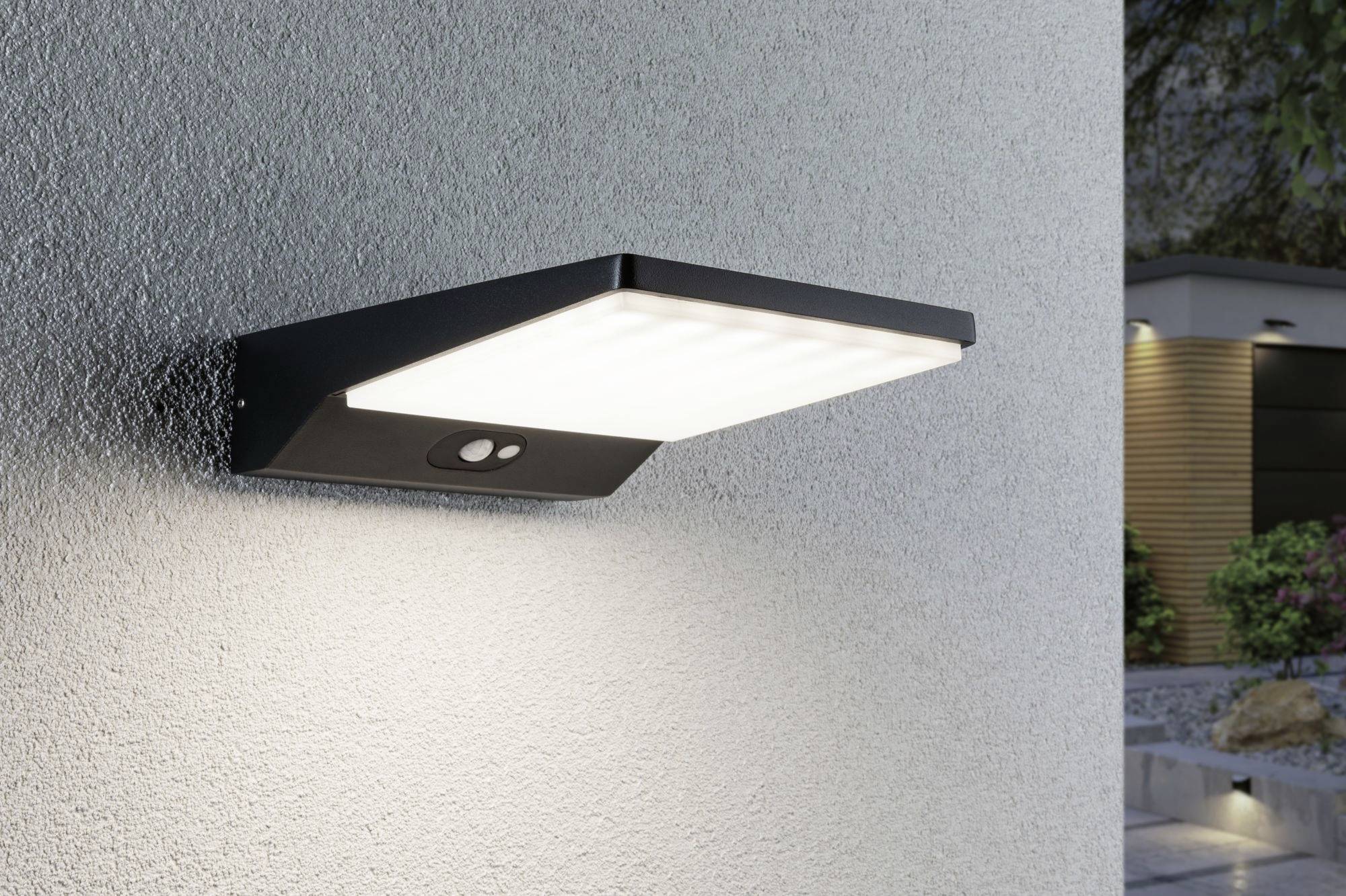 Nástěnná lampa s pohybovým senzorem na šedé venkovní zdi, svítící za soumraku. V pozadí zahradní prostor.