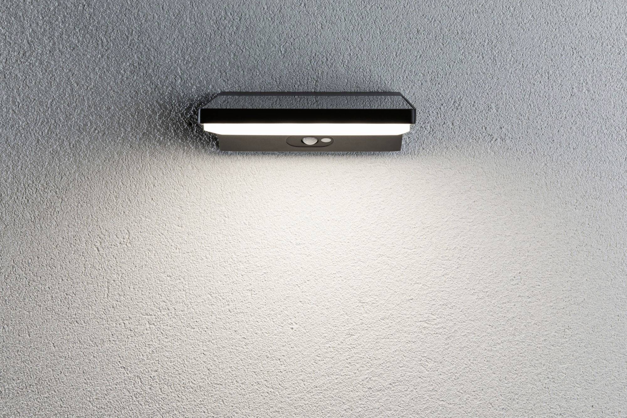 Nástěnná lampa s pohybovým senzorem svítí na šedé zdi a osvětluje prostor pod sebou.
