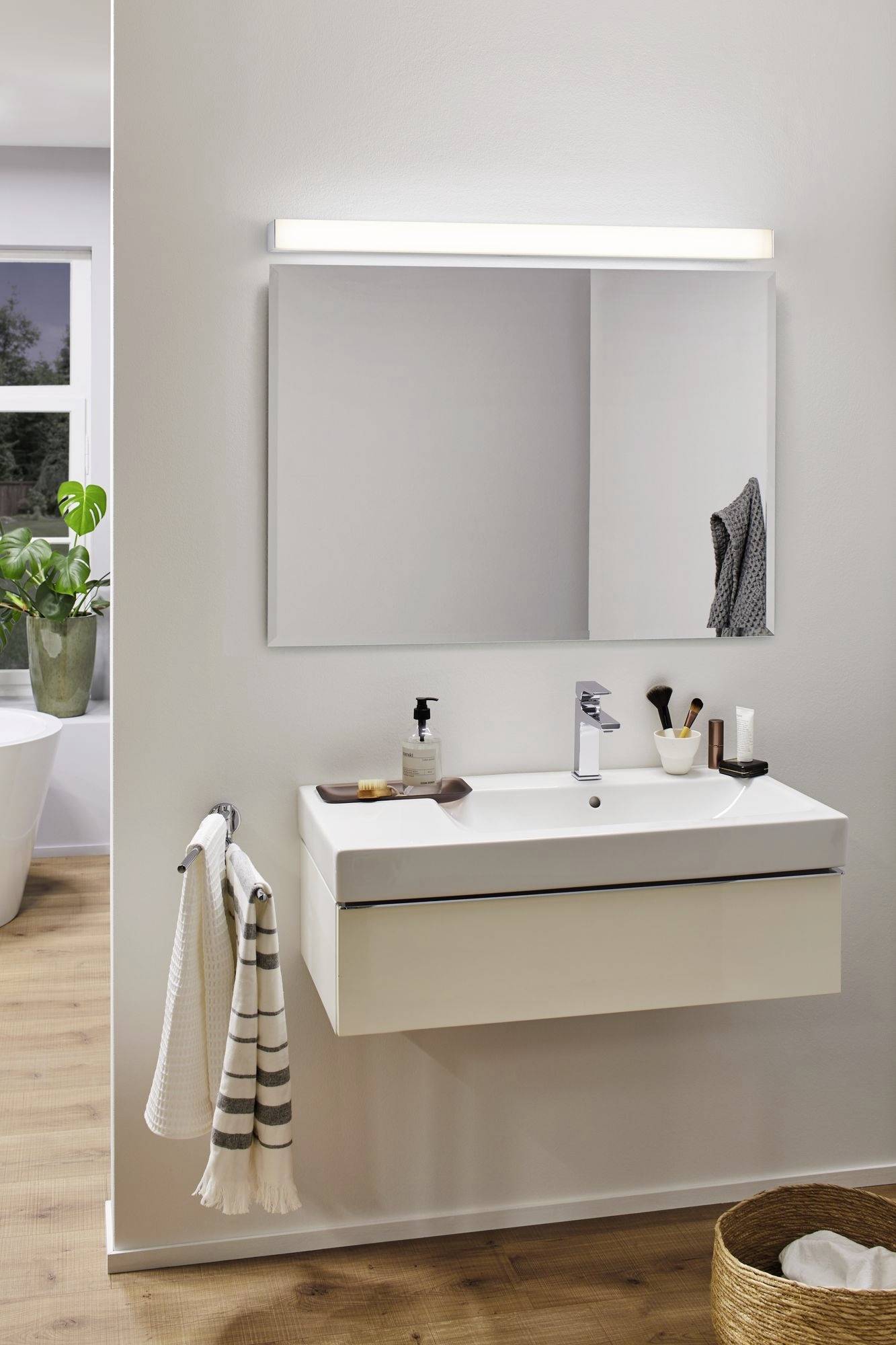 Moderní koupelna s umyvadlem, zrcadlem a světlou dřevěnou podlahou. Ručník a hygienické produkty na koupelnovém pultu.