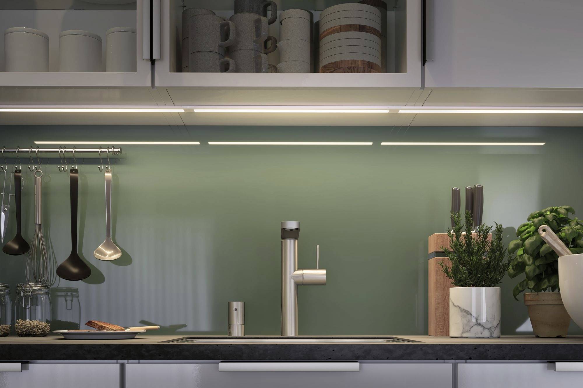 Moderní kuchyně se zelenohnědou stěnou, nerezovou dřezem a vařnou deskou. Kuchyňské náčiní visí na stěně vedle čerstvých bylinek.