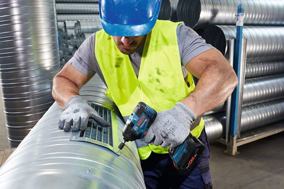 Dělník v ochranné přilbě a vestě upevňuje mříž na velké vzduchotechnické potrubí pomocí akumulátorového šroubováku v dílně.