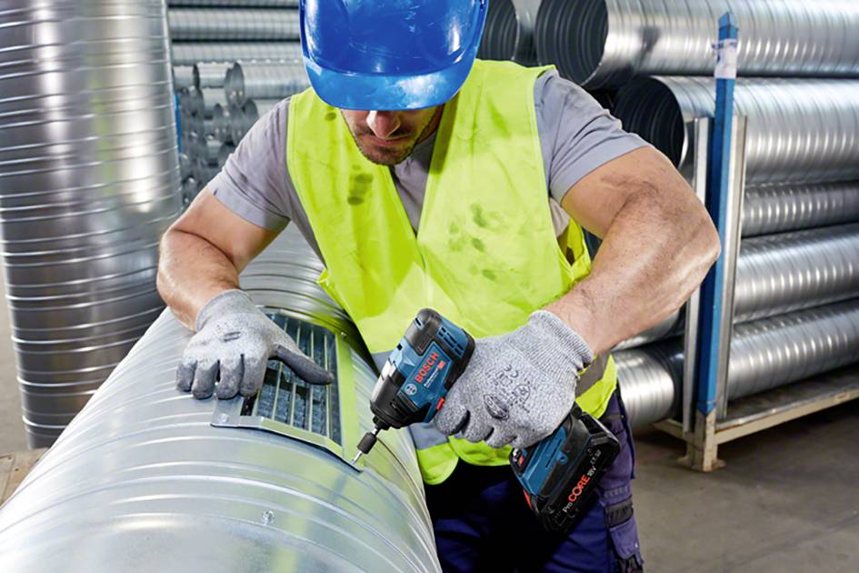Dělník v modré ochranné přilce a žluté vestě šroubuje akumulátorovým šroubovákem na velkém ventilačním potrubí v dílně.