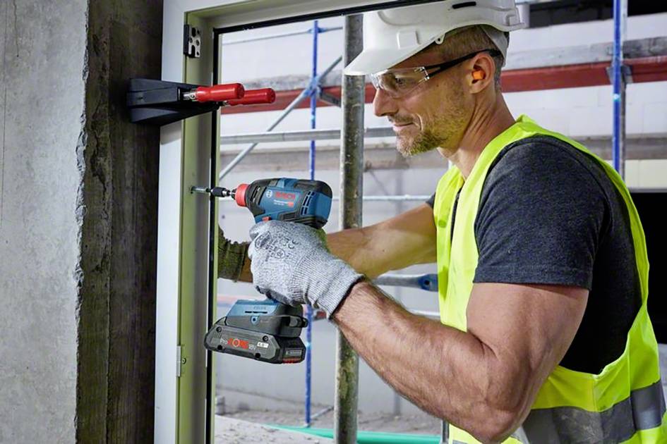 Dělník v ochranné přilbě a reflexní vestě montuje okenní rám pomocí vrtačky na budově.