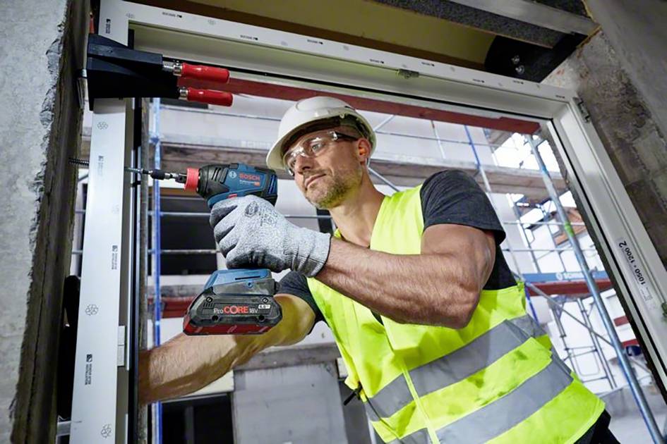 Dělník v helmě a reflexní vestě upevňuje okenní rám pomocí elektrické vrtačky na staveništi.